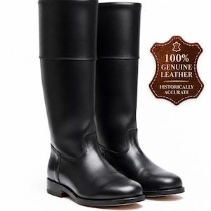 May include: A pair of black knee-high leather boots with a smooth finish, rounded toes, and low heels. A brown leather-shaped badge is present with the text 100% GENUINE LEATHER HISTORICALLY ACCURATE, featuring a star icon at the top center.