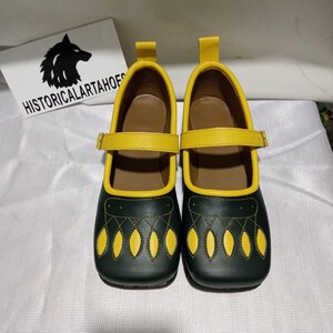 Op de afbeelding: Een paar donkergroene Mary Jane-schoenen met felgele randen, een geel enkelbandje met een metalen gesp en een geel treklipje bij de hiel. De neus is voorzien van decoratieve gele stiksels en uitsparingen. Een bordje toont HISTORICALARTASHOES.