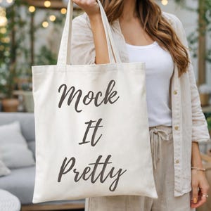 May include: A cream-colored canvas tote bag featuring the dark gray text Mock It Pretty printed in a cursive font on the front. The bag has two long shoulder straps and a rectangular shape, constructed from a woven fabric material.
