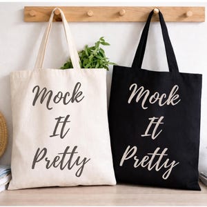 以下が含まれることがあります： 「Mock It Pretty」の文字が筆記体で書かれた、クリーム色と黒色の2つのトートバッグ。木製のラックに掛けられています。麦わら帽子、鉢植えの多肉植物、本が背景にあります。