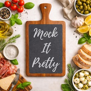 Puede incluir: Una tabla de cortar de madera con un centro de pizarra negra, con el texto "Mock It Pretty" en escritura blanca. La tabla está rodeada de varios alimentos, como tomates, aceitunas, pan y queso, lo que sugiere un tema culinario.