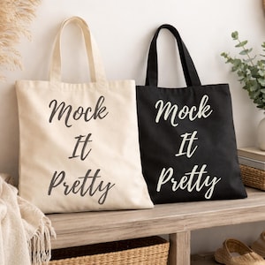 May include: Two tote bags, one cream and one black, with the phrase "Mock It Pretty" in white script. The bags have long handles and are made of a sturdy fabric. They are displayed on a wooden bench.