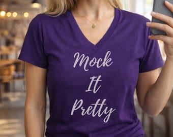 May include: A purple V-neck t-shirt with the words "Mock It Pretty" in a cursive font. The shirt is a solid color and appears to be made of a soft material. The model is wearing a gold necklace.