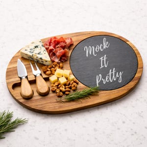 May include: Wooden cheese board with a dark slate insert, featuring the text "Mock It Pretty". The board holds cheese, nuts, prosciutto, and a sprig of rosemary. Two cheese knives are included.