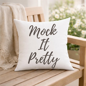 May include: A square, white decorative pillow with the words "Mock It Pretty" in dark gray script. The pillow is resting on a wooden bench with a cream-colored throw blanket in the background.