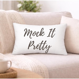 May include: A white rectangular cushion with the words "Mock It Pretty" in black script sits on a beige sofa. A textured throw and two other cushions in beige tones are also visible.