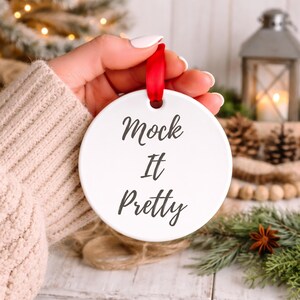 May include: A white, round Christmas ornament with the text "Mock It Pretty" in black script. The ornament has a red ribbon for hanging. The background includes a Christmas tree, lantern, and pinecones.