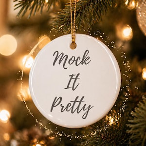 Puede incluir: Adorno navideño blanco y redondo con la frase "Mock It Pretty" en negro, con una fuente cursiva. El adorno cuelga de un hilo dorado sobre un fondo borroso de luces navideñas y vegetación.