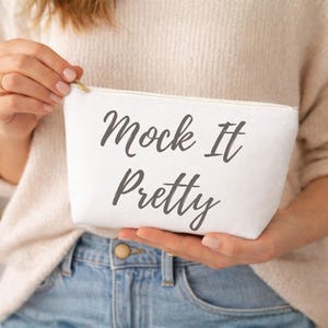 May include: A white canvas cosmetic bag with the text "Mock It Pretty" in a dark script font. The bag has a gold zipper and is held by a person wearing a cream-colored sweater and blue jeans.