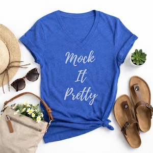 May include: A royal blue V-neck t-shirt with the text "Mock It Pretty" in a cursive font. The shirt is styled with sunglasses, a straw hat, a beige purse with flowers, and brown sandals.