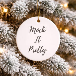 Op de afbeelding: Een witte, ronde kerstversiering met de tekst "Mock It Pretty" in zwart schrift. De versiering hangt aan een gouden draad tegen een achtergrond van een besneeuwde kerstboom.