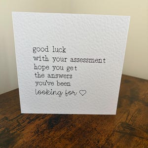 May include: A square white greeting card with a textured surface featuring black printed text that reads: good luck with your assessment hope you get the answers you've been looking for followed by a small hand-drawn heart symbol.