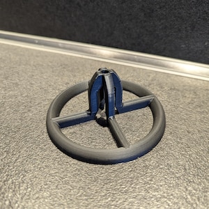 Könnte beinhalten: Ein dunkelgraues, 3D-gedrucktes Kunststoffteil. Das Teil hat eine kreisförmige Basis mit einem kreuzförmigen Design in der Mitte. Eine vertikale, geschlitzte Struktur erhebt sich aus der Mitte des Kreuzes.