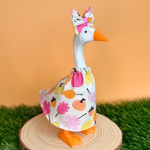 May include: A white plastic goose figurine wearing a fabric dress with a pattern of colorful autumn leaves, pumpkins, and acorns. The goose has an orange beak and feet, and wears a matching fabric bow on its head with a bright pink center band.