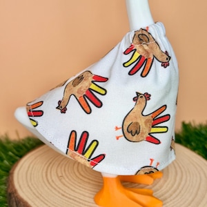 May include: A white fabric skirt featuring a repeating pattern of brown turkeys with colorful red, orange, and yellow tail feathers. The skirt has an elasticized waistband and is worn by a white goose figurine with bright orange webbed feet.