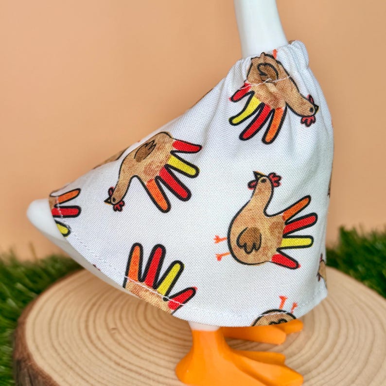 May include: A white fabric skirt featuring a repeating pattern of brown turkeys with colorful red, orange, and yellow tail feathers. The skirt has an elasticized waistband and is worn by a white goose figurine with bright orange webbed feet.
