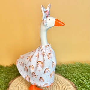 May include: A white goose figurine with an orange beak and feet, wearing a patterned skirt and a matching headpiece. The skirt and headpiece feature a repeating rainbow design in shades of brown and pink. The goose is posed on a wooden slice.