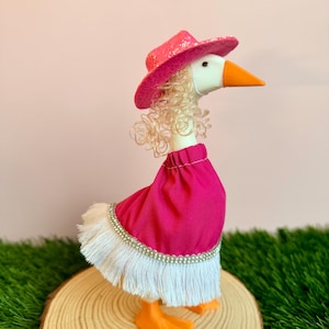 May include: A white goose figurine wearing a bright pink glittery cowboy hat, blonde curly hair, and a vibrant pink skirt featuring a white fringe trim with a silver sequin border. The goose has an orange beak and stands on orange feet.