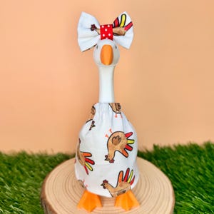 May include: A white goose figurine wearing a white fabric skirt featuring a repeating pattern of brown and orange chickens. The goose has a bright orange beak and is adorned with a white hair bow featuring red polka dots and a central red band.