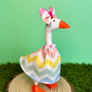 May include: A whimsical goose figurine with a white body, orange beak and feet, and a pink and white striped bow. The goose wears a skirt with a colorful chevron pattern in pink, yellow, and blue. The figurine is displayed on a wooden slice.