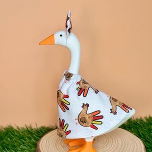 May include: A white goose figurine wearing a white fabric dress featuring a repeating pattern of brown handprint turkeys with orange, yellow, and red fingers. The goose has an orange beak and a matching fabric headband with a small bow on top.