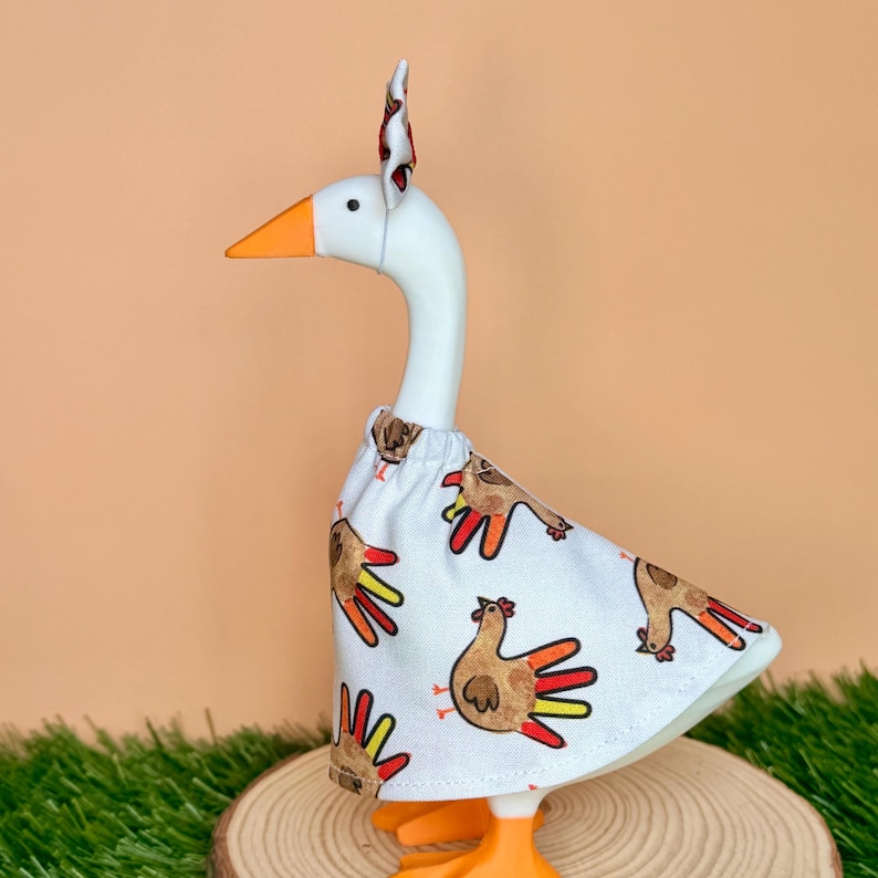 May include: A white goose figurine wearing a white fabric dress featuring a repeating pattern of brown handprint turkeys with orange, yellow, and red fingers. The goose has an orange beak and a matching fabric headband with a small bow on top.
