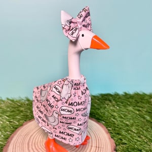 May include: A decorative goose figurine with a pink body, orange beak and feet, and a pink bow. The goose wears a pink dress with a repeating pattern of the word "MOM?" and a cartoon bird illustration. The figurine is displayed on a wooden slice.