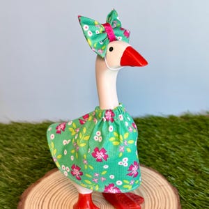 May include: A whimsical goose figurine with a white body, red beak and feet, and a green floral dress and bow. The bow is accented with a pink center. The goose is standing on a wooden slice, creating a charming decorative piece.