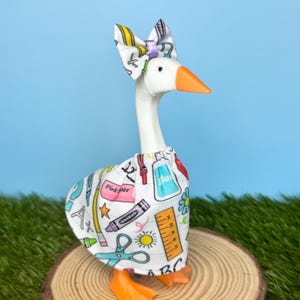 May include: A white goose figurine wearing a fabric skirt and a matching bow on its head. The fabric features a colorful school-themed pattern with crayons, rulers, scissors, glue, and the letters ABC on a white background. The goose has an orange beak.