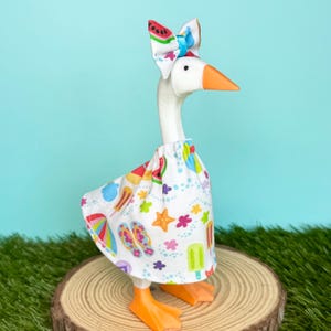 May include: A white goose figurine wearing a white fabric dress featuring a colorful summer-themed pattern of watermelons, popsicles, flip-flops, and starfish. The goose has an orange beak and a matching fabric bow on its head with a blue ribbon.