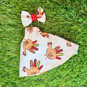 May include: A cream-colored fabric cone-shaped accessory featuring a repeating print of brown chickens with red and yellow tail feathers. The top is adorned with a small cream bow featuring a central red band with white polka dots.