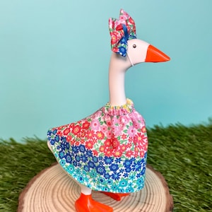 May include: A whimsical goose figurine adorned with a floral print dress and matching bow. The goose has an orange beak and feet, and a white face. The dress features a vibrant mix of red, pink, blue, and green flowers. The figurine is displayed on a wooden slice.