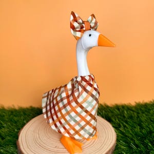 May include: A white goose figurine wearing a brown, orange, and cream plaid fabric dress and a matching plaid bow on its head. The goose has a bright orange beak and orange feet, standing upright with a smooth, glossy finish on its neck and head.