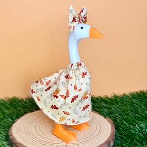 May include: A white goose figurine wearing a cream-colored dress with a pattern of red, orange, and brown autumn leaves. The goose has an orange beak and feet, and a matching fabric bow is attached to the top of its head between its eyes.