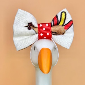 May include: A white fabric bow with a turkey print featuring red, yellow, and brown feathers. The center of the bow is a red ribbon with white polka dots. The bow is attached to a white goose figurine with a bright orange beak and eyes.