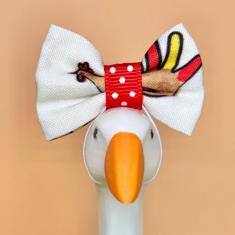 May include: A white fabric bow with a turkey print featuring red, yellow, and brown feathers. The center of the bow is a red ribbon with white polka dots. The bow is attached to a white goose figurine with a bright orange beak and eyes.