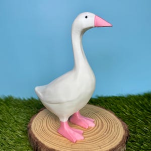 May include: A white goose figurine with a pink beak and pink webbed feet. The goose is standing in a profile position with its neck extended upward, featuring a smooth surface and a small black dot for an eye, captured in a clear, bright view.