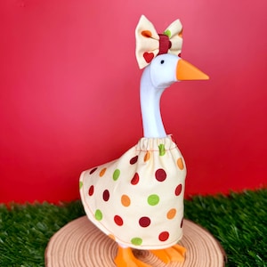 May include: A white goose figurine wearing a cream-colored fabric dress with a pattern of red, orange, and green polka dots. The goose has an orange beak and a matching cream-colored bow on its head featuring red hearts and a dark red center band.