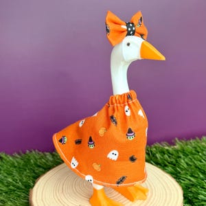 May include: A white plastic goose figurine wearing an orange fabric dress featuring a pattern of ghosts, pumpkins, bats, and witch hats. The goose has an orange beak and an orange bow with black and white polka dots attached to its head.