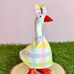 May include: A whimsical goose figurine with a white body, red feet and beak, and a pastel plaid skirt and bow. The goose is standing on a wooden slice, with a pink background and green grass.