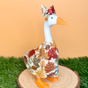 May include: A white goose figurine wearing a dress featuring a pattern of autumn leaves in shades of orange, brown, yellow, and green. The goose has an orange beak and feet, and wears a matching bow on its head with a small red ribbon detail.