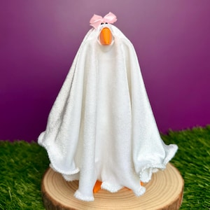 May include: A white fabric-covered goose figurine featuring a small pink bow on its head and a bright orange beak. The goose is draped in a white cloth that hangs down to the base, creating a ghost-like appearance with visible orange feet.