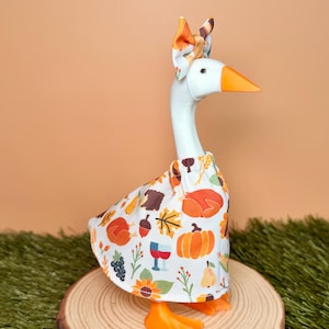 May include: A white plastic goose figurine wearing a fabric dress with a colorful autumn-themed print featuring pumpkins, turkeys, grapes, and acorns. The goose has an orange beak and feet, and wears a matching fabric bow on its head.