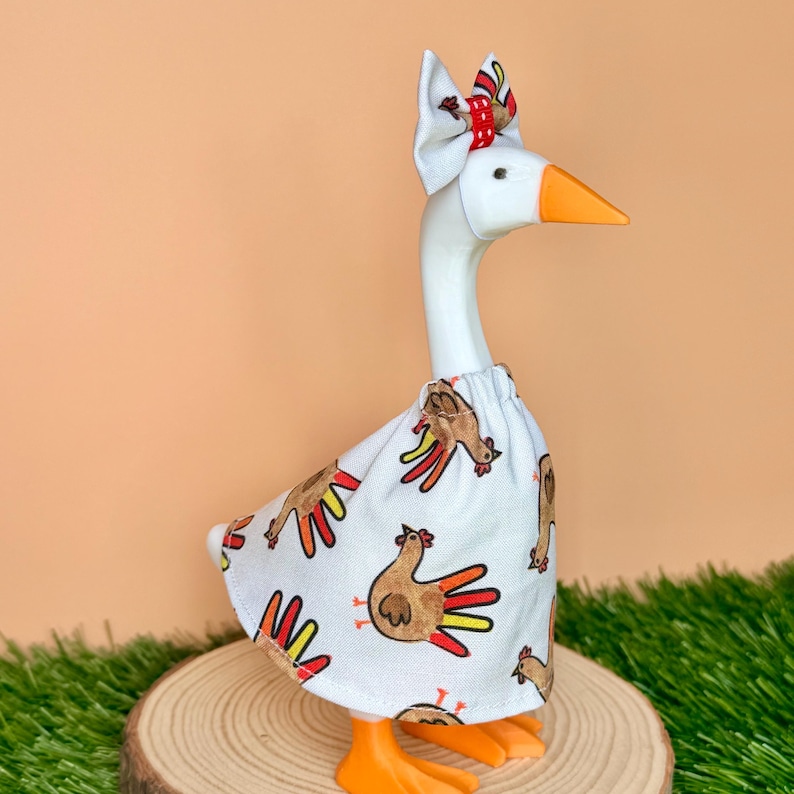 May include: A white goose figurine wearing a white fabric dress featuring a repeating pattern of brown turkeys with red and yellow tail feathers. The goose has an orange beak and feet, and a matching white fabric bow with a red center on its head.