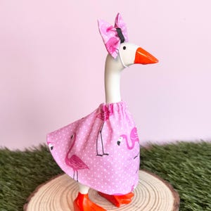 May include: A white goose figurine wearing a pink dress with a white polka dot and flamingo pattern. The goose has an orange beak and a matching pink polka dot bow attached to its head with a black band. The figurine stands on orange feet.