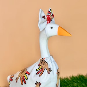 May include: A white goose figurine wearing a white fabric outfit featuring a repeating turkey pattern with red, yellow, and brown colors. The goose has an orange beak and a small white bow on its head with a red and white polka dot center accent.