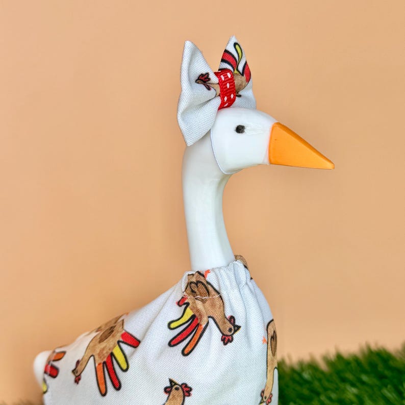 May include: A white goose figurine wearing a white fabric outfit featuring a repeating turkey pattern with red, yellow, and brown colors. The goose has an orange beak and a small white bow on its head with a red and white polka dot center accent.