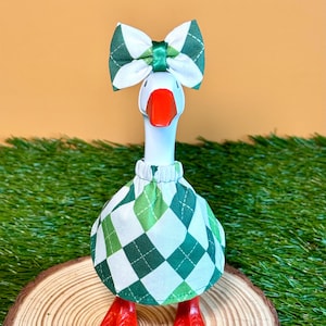 May include: A decorative goose figurine with a white body, red feet and beak, and a green and white argyle patterned dress and bow. The goose is standing on a wooden slice against a backdrop of green grass and a yellow-orange background.