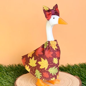 May include: A white plastic goose figurine wearing a fabric dress and a matching bow on its head. The dress and bow feature a repeating pattern of autumn leaves in shades of red, orange, yellow, and green against a dark brown background.