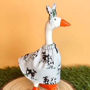 May include: A decorative goose figurine wearing a cow-print skirt and matching bow. The goose has an orange beak and feet, with a white body and neck. The skirt and bow feature a light gray background with black and white cow illustrations.